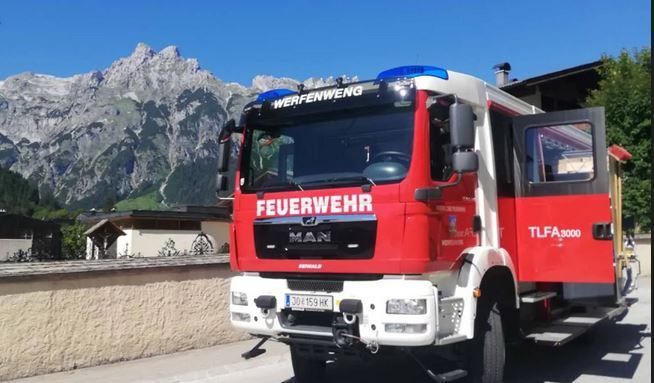 Ein roter und weißer Feuerwehrwagen mit der Aufschrift 'Feuerwehr' und 'MAN' mit einer blauen Licht auf der Oberseite ist vor einem Gebäude geparkt, mit einer Bergkette im Hintergrund.