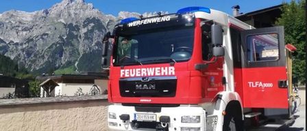 Ein roter und weißer Feuerwehrwagen mit der Aufschrift 'Feuerwehr' und 'MAN' mit einer blauen Licht auf der Oberseite ist vor einem Gebäude geparkt, mit einer Bergkette im Hintergrund.