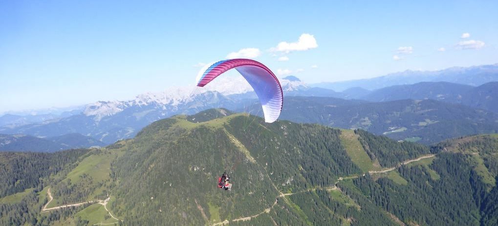 Eine Person paragliding über einer Berglandschaft mit einem rosa und blauen Fallschirm gegen einen blauen Himmel und weiße Wolken.