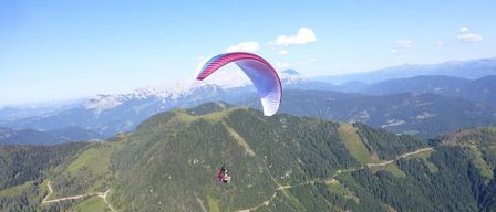 Eine Person paragliding über einer Berglandschaft mit einem rosa und blauen Fallschirm gegen einen blauen Himmel und weiße Wolken.