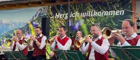 Eine Gruppe von Musikern in traditioneller bayerischer Kleidung spielt Trompeten unter einem Zelt mit einer malerischen Kulisse. Das Banner sagt 'Herzlich willkommen'.