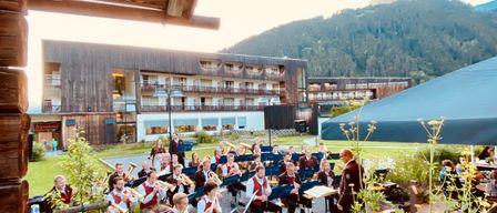 Ein Blasorchester spielt auf einer Rasenfläche mit Gebäude und Bergen im Hintergrund. Die Musiker sitzen mit ihren Instrumenten auf Bänken.