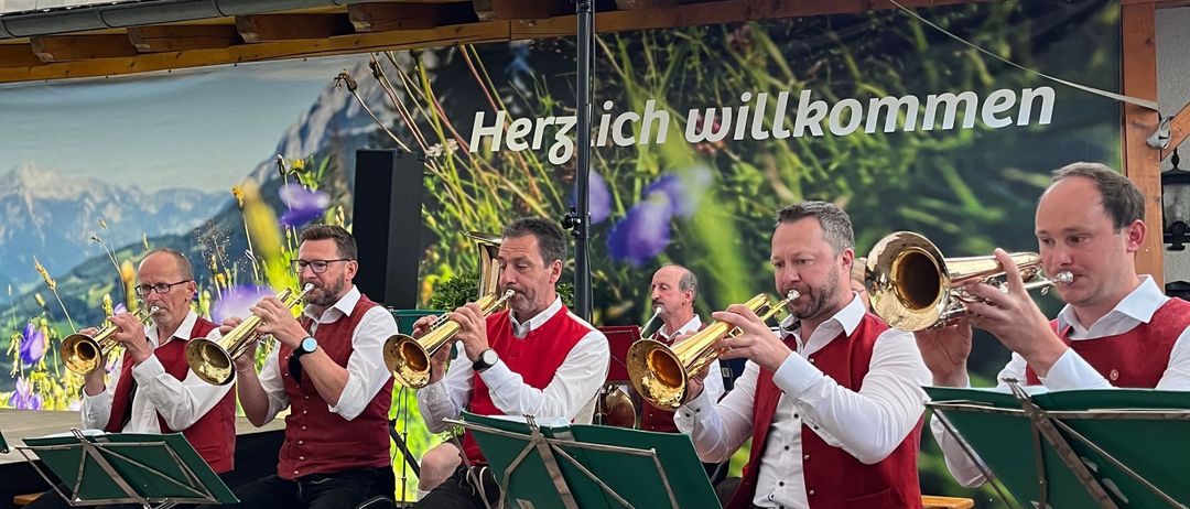 Fünf Männer spielen Trompeten auf einer Bühne. Sie tragen traditionelle Kostüme. Hinter ihnen ist eine Kulisse mit einer Bergkette und den Worten 'Herz ich willkommen'.
