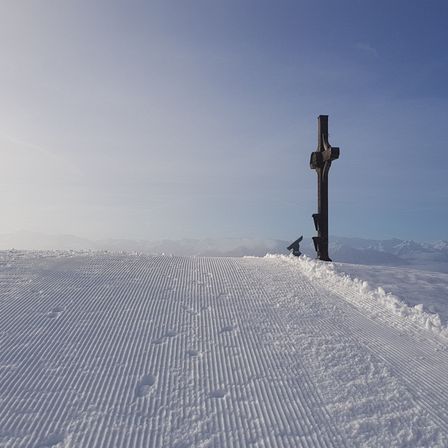 Ein Kreuz steht auf einem verschneiten Hang mit einer Skilift-Anlage im Hintergrund. Der Himmel ist klar und blau.