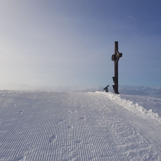 Ein Kreuz steht auf einem verschneiten Hang mit einer Skilift-Anlage im Hintergrund. Der Himmel ist klar und blau.