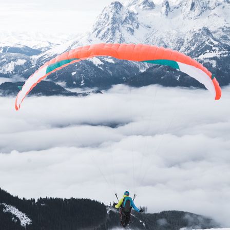 Eine Person paragliding mit einem orangefarbenen und türkisfarbenen Fallschirm fliegt über Wolken mit verschneiten Bergen im Hintergrund.