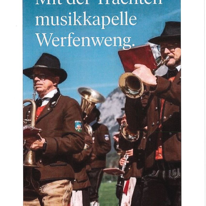 Eine Gruppe von Musikern in traditioneller Kleidung spielt im Freien. Sie halten Blechblasinstrumente. Ein Mann vorne hält ein Buch. Hinter ihnen ist ein verschwommener Hintergrund von Bergen.