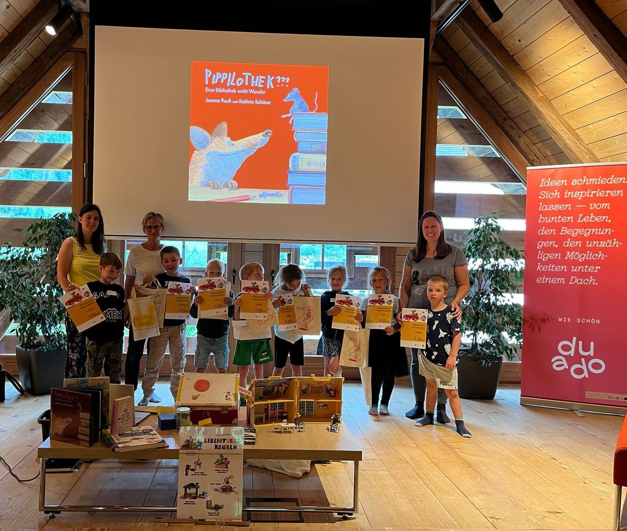 Mehrere Kinder halten Bücher und Zertifikate in einem Raum mit einer Projektionsleinwand, Erwachsene stehen hinter ihnen und ein Banner mit Logo und Text ist rechts.