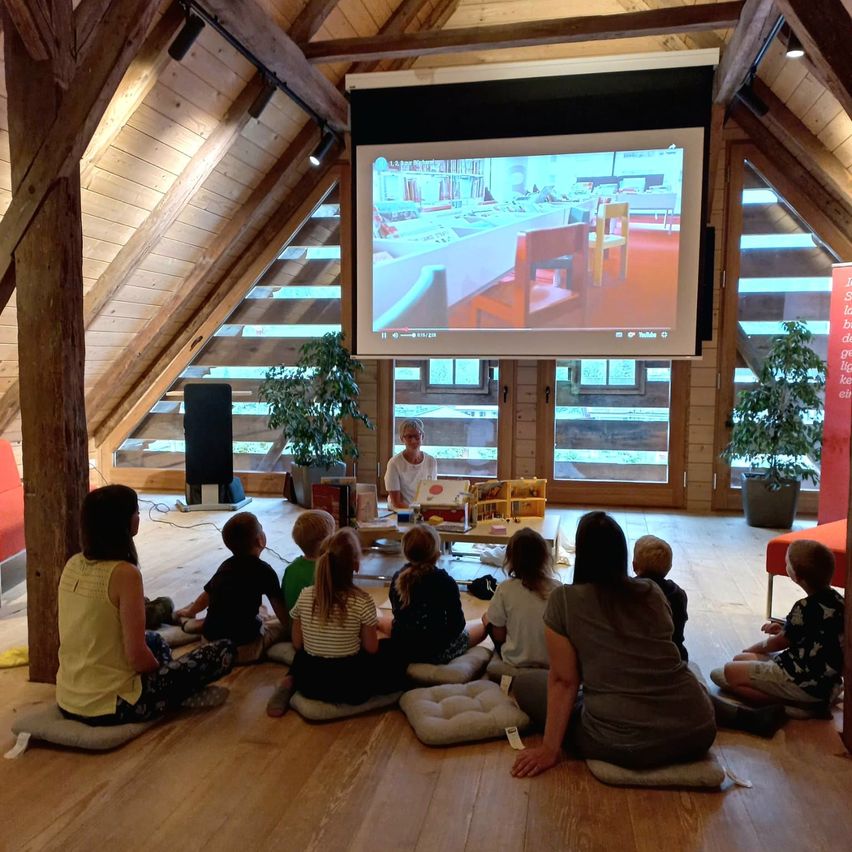 Kinder und Erwachsene sitzen auf Kissen in einem Dachzimmer und schauen auf eine Projektionsleinwand an der Decke. Ein Mann steht an einem Tisch mit Büchern. Zwei Topfpflanzen stehen neben den Türen.