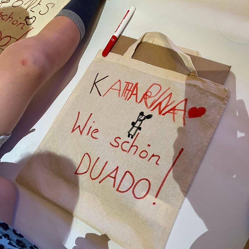 Ein Stoff-Tragetasche mit rotem Text, der 'KATHARINA Wie schön DUADO!' mit einem Herzsymbol und einem Panda-Zeichnung liest. Ein weißer Marker befindet sich neben der Tasche.