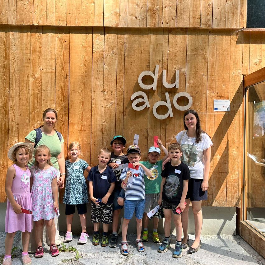 Eine Gruppe von Kindern und Erwachsenen steht vor einer Holzwand mit der Aufschrift 'du aDo'. Sie lächeln und halten rote Fahnen.