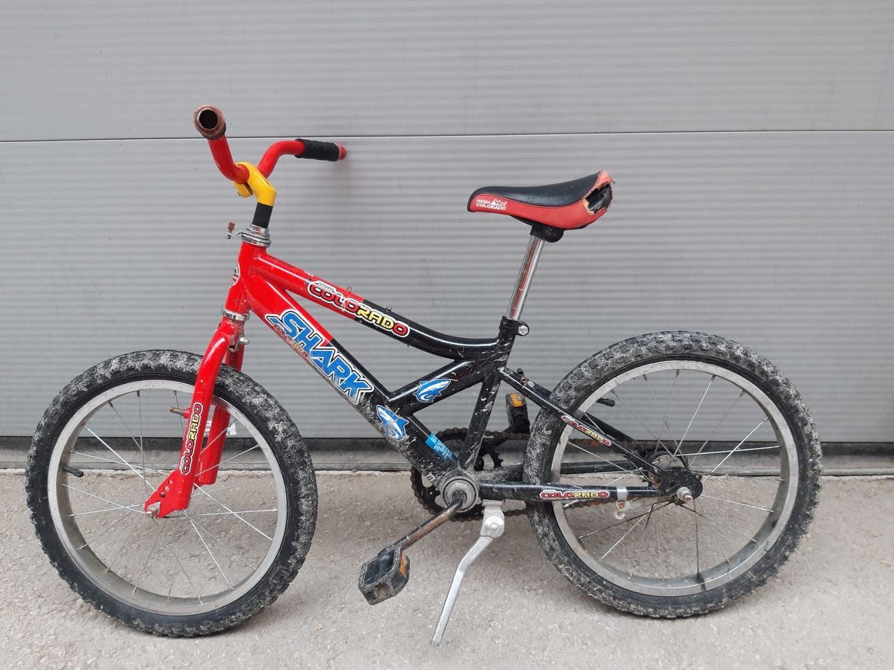 Ein rotes und schwarzes Fahrrad mit dem Wort Shark auf der Seite. Das Fahrrad hat einen beschädigten Sitz und steht auf dem Boden.