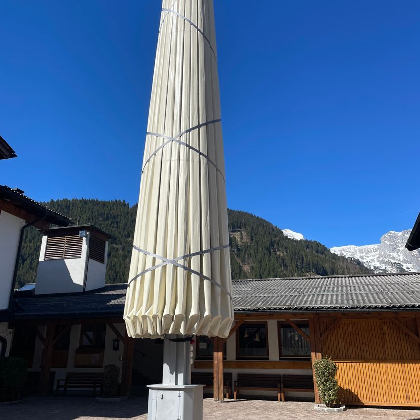 A large outdoor umbrella is rolled up and secured with straps against a backdrop of mountains. A wooden building with a gray roof is visible in the foreground.