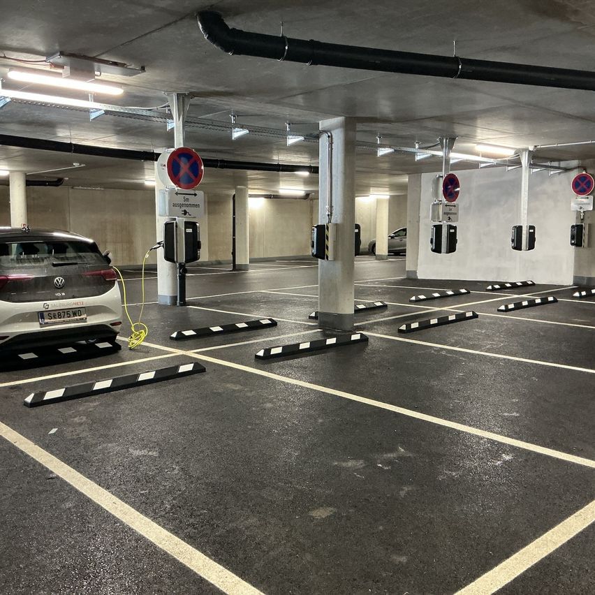 Eine Tiefgarage mit einer Elektroauto-Ladestation. Das Auto ist geparkt und lädt. Die Tiefgarage hat weiß-schwarz gestreifte Barrieren.