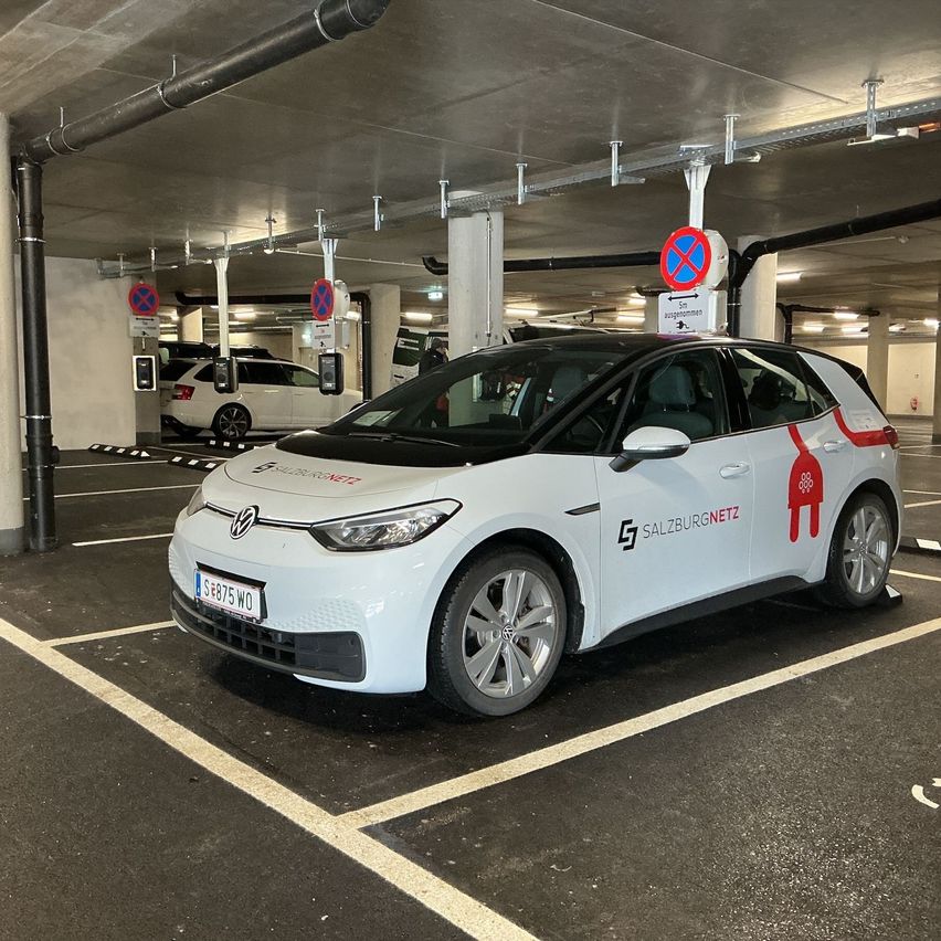Ein weißer Elektrowagen mit dem Logo Salzburgnetz steht in einer Garage. Dahinter ist ein weißer SUV geparkt. Schilder sind an der Decke und an der Säule angebracht.