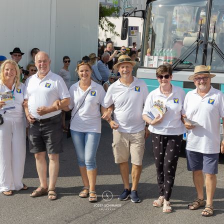 Eine Gruppe von Menschen, die gleiche weiße Hemden tragen, steht vor einem Bus und lächelt für die Kamera. Sie halten verschiedene Broschüren in den Händen. Dahinter sind weitere Menschen versammelt, und ein Bus ist zu sehen.