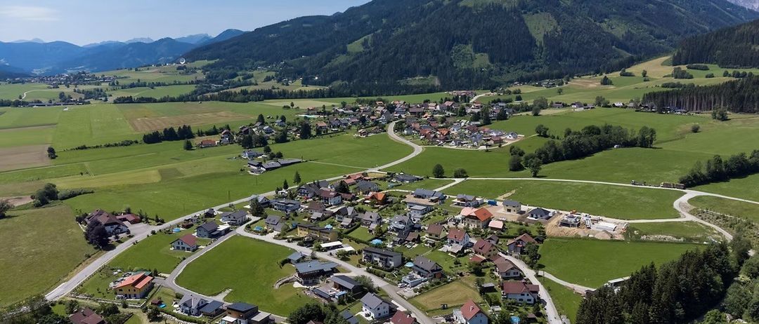 Luftaufnahme eines kleinen Dorfes, umgeben von satten grünen Feldern und bewaldeten Bergen in der Ferne.