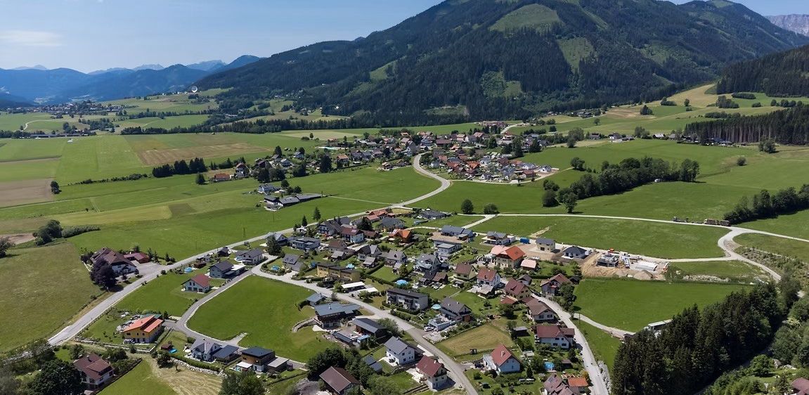 Luftaufnahme eines kleinen Dorfes, umgeben von satten grünen Feldern und bewaldeten Bergen in der Ferne.