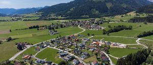 Luftaufnahme eines kleinen Dorfes, umgeben von satten grünen Feldern und bewaldeten Bergen in der Ferne.