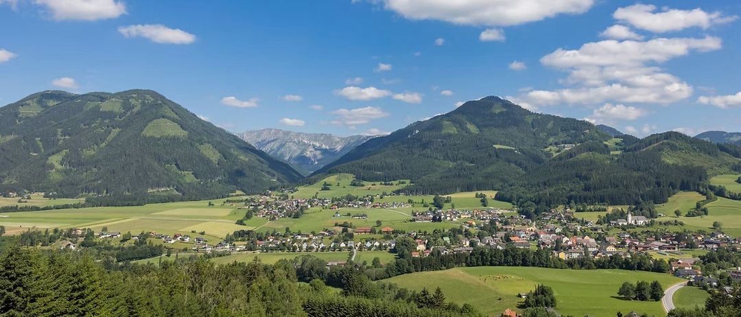 Ein Panoramablick auf ein Tal mit einem kleinen Dorf, umgeben von üppigen grünen Feldern, Bäumen und Bergen unter einem blauen Himmel mit Wolken.
