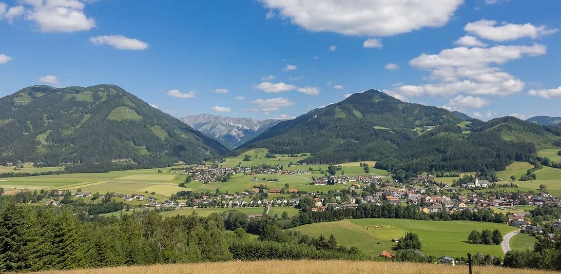 Ein Panoramablick auf ein Tal mit einem kleinen Dorf, umgeben von üppigen grünen Feldern, Bäumen und Bergen unter einem blauen Himmel mit Wolken.