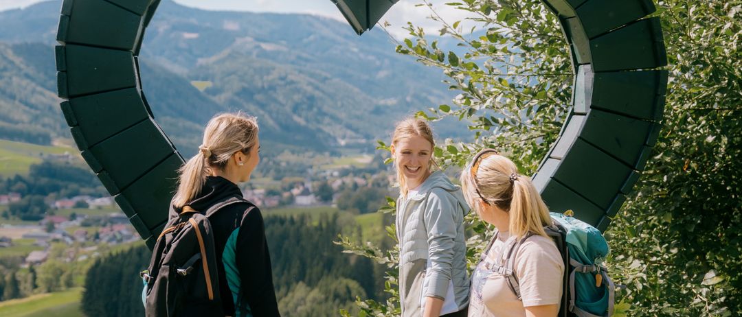 Drei Frauen stehen in der Nähe einer grünen Herzskulptur auf einem Berg mit Blick auf entfernte Häuser und Berge.