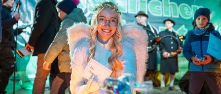Eine Frau in einem Engelkostüm lächelt und hält eine Karte, umgeben von Menschen in Winterkleidung, mit einem Banner im Hintergrund.