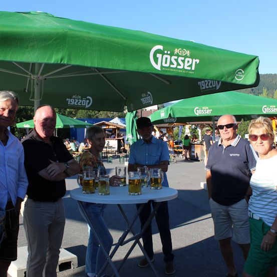 Eine Gruppe von Menschen steht unter einem grünen Gosser-Schirm, mit Gläsern Bier auf einem Tisch vor ihnen.