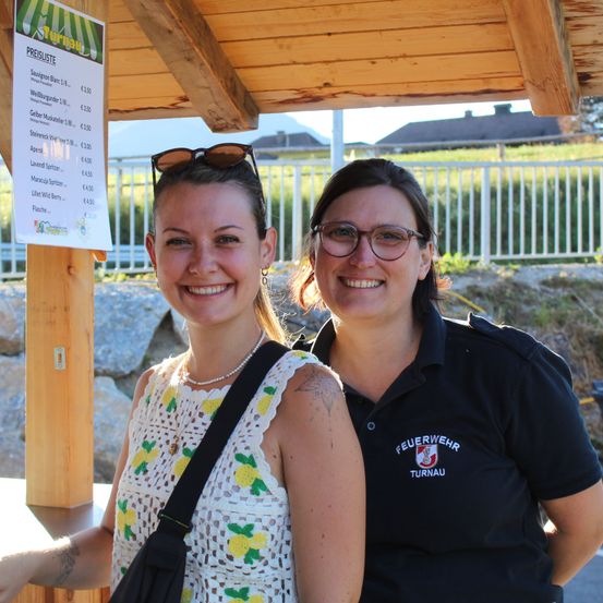 Zwei Frauen stehen lächelnd unter einem hölzernen Pavillon mit einer Preisliste für Getränke auf der linken Seite. Die Frau auf der linken Seite trägt ein weißes Blumenoberteil und eine Brille auf dem Kopf. Die Frau auf der rechten Seite trägt ein schwarzes Shirt mit dem Wort 'Feuerwehr' darauf.