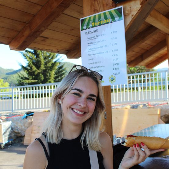Eine Frau mit blonden Haaren, die ein schwarzes Oberteil trägt und einen Hot Dog in einem Imbissstand hält, mit einer Preisliste im Hintergrund.