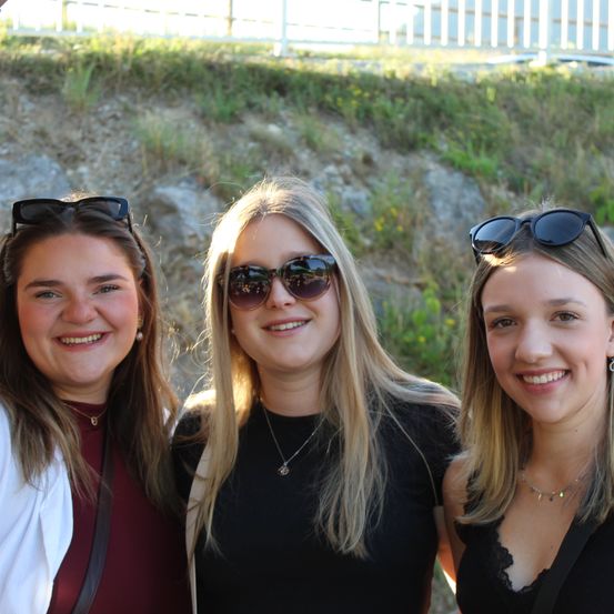 Drei Frauen stehen lächelnd nebeneinander vor einem Grashügel. Sie tragen alle Sonnenbrillen auf dem Kopf.