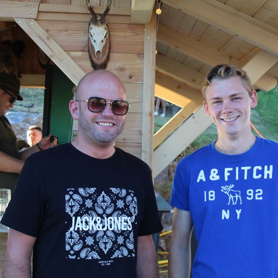 Zwei Männer stehen nebeneinander, einer trägt ein schwarzes Jack & Jones-Shirt und der andere ein blaues A&Fitch-Shirt, lächeln vor einer Holzstruktur mit einem aufgehängten Hirschschädel an der Wand.
