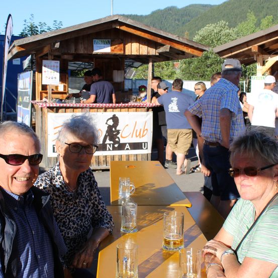 Drei Personen sitzen an einem Tisch mit Gläsern Bier vor ihnen. Dahinter steht ein kleiner Holzstand mit einem Banner, auf dem 'Club' steht. Mehrere Personen stehen herum, einige tragen Hüte und Sonnenbrillen. Im Hintergrund sind Berge und Bäume zu sehen.