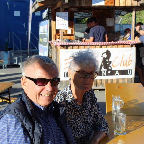 Ein älteres Paar sitzt an einem Tisch in einer Außenumgebung, lächelt und posiert für ein Foto. Hinter ihnen ist ein hölzerner Kiosk mit einem Banner zu sehen, das 'Club N A U' liest.