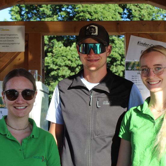 Drei Personen in grünen Poloshirts und Sonnenbrillen lächeln für ein Foto vor einer Holzstruktur mit Schildern und Bäumen im Hintergrund.