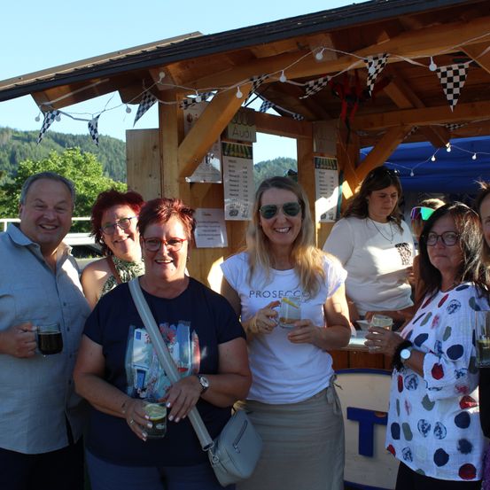 Eine Gruppe von Frauen und Männern steht unter einem hölzernen Pavillon, lächelt und hält Getränke. Sie sind lässig gekleidet und scheinen sich bei einer sozialen Veranstaltung zu befinden.