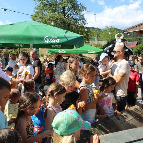 Eine Gruppe von Kindern und Erwachsenen versammelt sich um einen Tisch, einige halten gelbe Objekte, möglicherweise auf einem Festival. Im Hintergrund sind grüne Zelte mit dem Wort Gosser zu sehen.