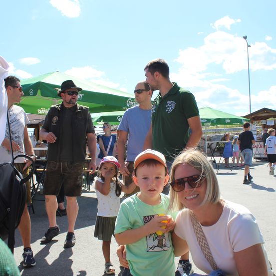 Eine vierköpfige Familie, einschließlich zweier Kinder, steht unter einem grünen Schirm in einem öffentlichen Bereich. Der Mann trägt einen Hut und die Frau eine Sonnenbrille. Die Kinder halten ein gelbes Objekt, möglicherweise ein Spielzeug.