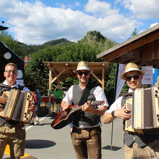Drei Männer in traditioneller bayerischer Kleidung, einer spielt Akkordeon, ein anderer Gitarre und der dritte spielt ebenfalls Akkordeon, stehen vor einer Holzstruktur mit einem Berg im Hintergrund.