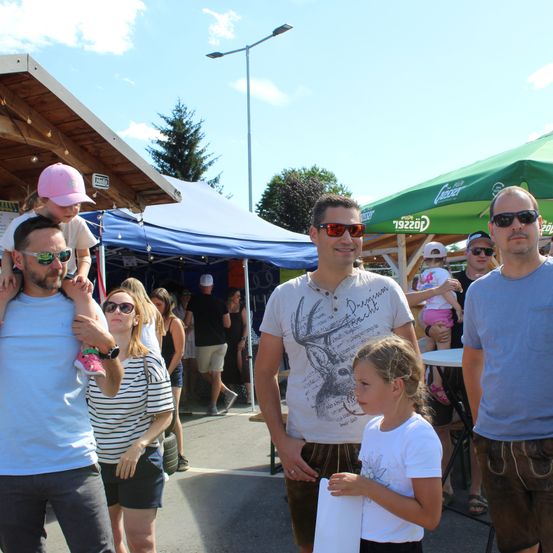 Eine Gruppe von Menschen, einschließlich Erwachsener und Kindern, steht in einem Außenbereich mit Zelten und einem klaren Himmel. Der Mann in der Mitte trägt eine Sonnenbrille und ein Hirsch-Print-Shirt.