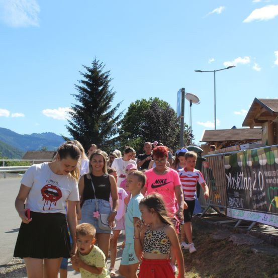 Eine Gruppe von Menschen, einschließlich Kindern, geht auf einem Pfad unter einem klaren blauen Himmel. Eine Frau in einem weißen Shirt mit roten Lippen führt die Gruppe an. Hinter ihnen ist ein Banner an einem Zaun befestigt.