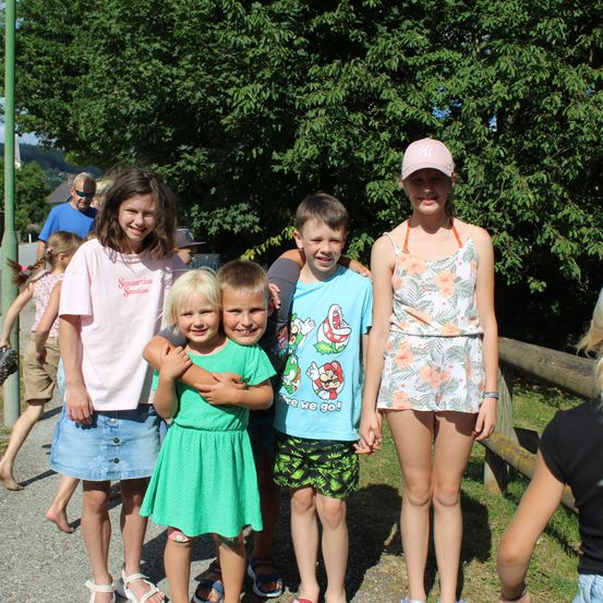 Fünf Kinder stehen an einem sonnigen Tag zusammen, mit einer Frau in einem rosa T-Shirt und einem Mädchen in einem Blumenkleid im Vordergrund. Dahinter stehen ein Junge in einem blauen T-Shirt mit einem Mario-Charakter und ein Junge in einem schwarzen Hut. Bäume und ein Holzzaun sind im Hintergrund zu sehen.