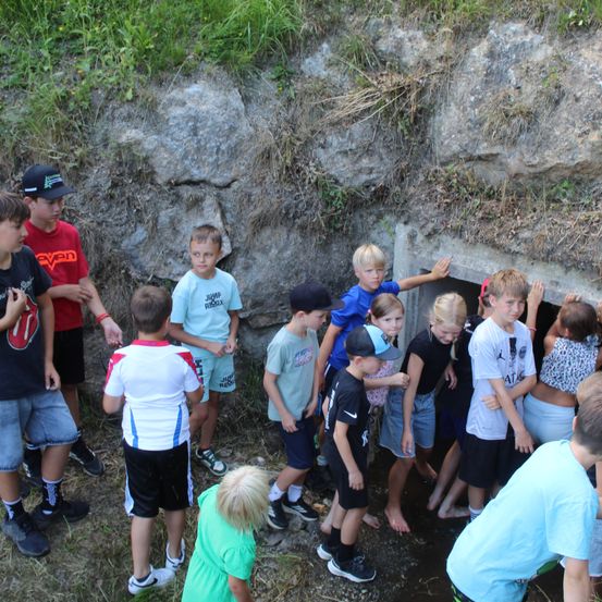 Eine Gruppe von Kindern steht um einen kleinen Wassergraben in der Nähe einer felsigen Wand. Einige sind barfuß, andere tragen Turnschuhe.