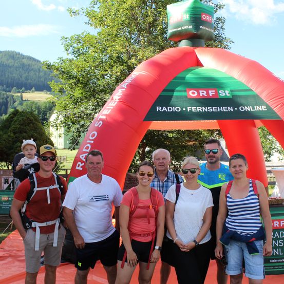 Eine Gruppe von Menschen steht unter einem aufblasbaren Bogen mit der Aufschrift 'Radio Steiermark' und 'ORF St', mit einem Berg im Hintergrund.