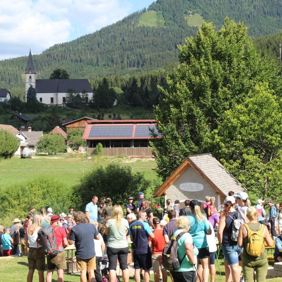 Eine Gruppe von Menschen steht auf einer grasigen Wiese mit einem kleinen Haus und einer Kirche im Hintergrund. Die Kirche hat einen Turm und ist von Bäumen umgeben. Es gibt ein Gebäude mit Solarmodulen auf dem Dach.