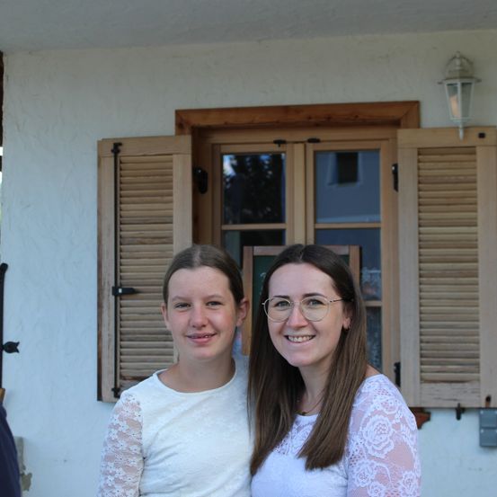 Zwei Frauen stehen vor einem Haus, lächeln und posieren für ein Foto. Die Frau auf der linken Seite trägt ein weißes Oberteil mit Spitzenärmeln. Die Frau auf der rechten Seite trägt eine Brille und ein weißes Oberteil. Hinter ihnen ist ein Fenster mit Holzläden.