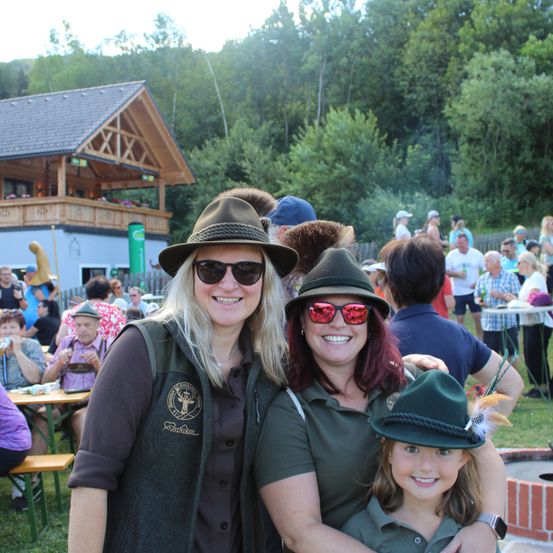 Drei Frauen in passenden grünen Hüten und Westen posieren für ein Foto bei einer Outdoor-Veranstaltung, mit einem Haus und Bäumen im Hintergrund.