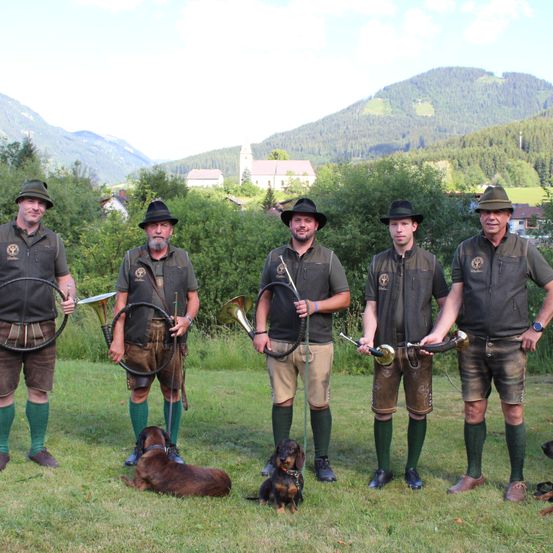 Fünf Männer in traditioneller bayerischer Kleidung, einschließlich grüner Socken und Hüten, stehen auf einem Rasen mit Bergen im Hintergrund. Sie halten Musikinstrumente und haben zwei Hunde zu ihren Füßen.