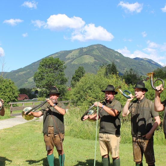 Vier Männer in traditioneller Kleidung spielen Blechblasinstrumente auf einer Wiese mit Bergen im Hintergrund.
