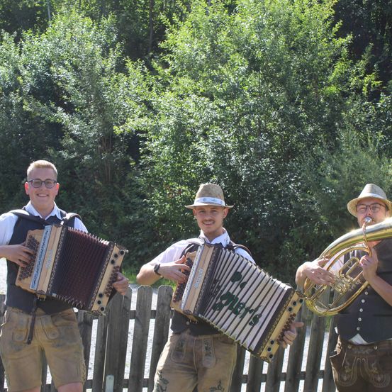 Drei Männer in traditioneller bayerischer Kleidung, einer spielt Akkordeon, ein anderer spielt eine französische Horn, und der dritte spielt ein Akkordeon, stehen vor einem Holzzaun mit Bäumen im Hintergrund.